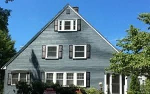 A charming, triangular blue house with white trim and dark shutters under a clear blue sky. Lush greenery surrounds the front entrance.