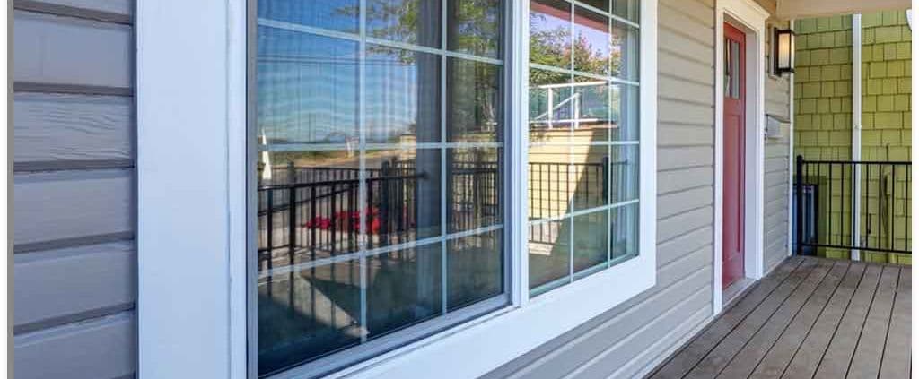 A wooden porch with light gray siding features a large window reflecting a railing. A red door and wall lamp are seen, creating a welcoming and tidy appearance.