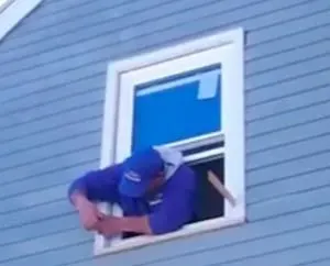 A person leans out of a small window in a blue clapboard house, attempting to install a window air conditioner. They wear a blue cap and hoodie, conveying a focused effort.