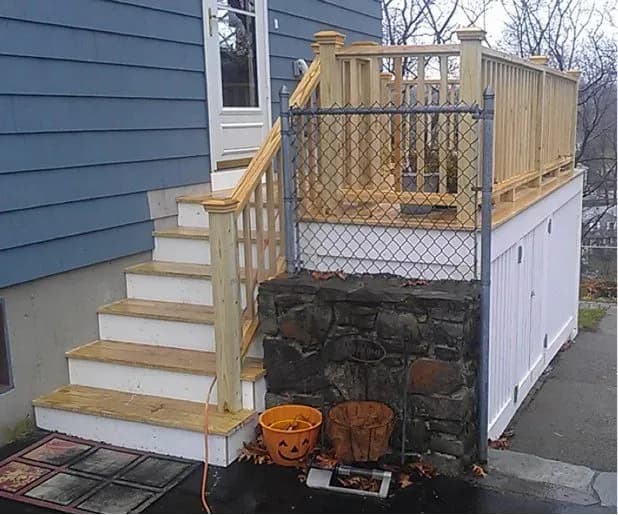 Wooden porch with a railing and steps leading up to a blue house. A decorative pumpkin pot and a rusty container sit on the ground, conveying an autumnal feel.