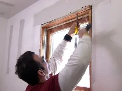 Man measuring a window with a screwdriver and hammer in a partially finished room. He wears safety goggles and a face mask, focused expression.