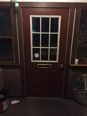 A wooden door with a grid-patterned window and a mail slot, flanked by dark-framed windows. The setting is dimly lit with a rustic, cozy ambiance.