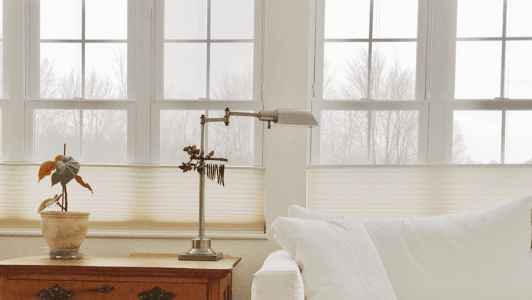 A cozy, minimalist room features large windows with soft light streaming in. A white sofa, wooden side table, potted plant, and a sleek lamp create a calm, serene atmosphere.