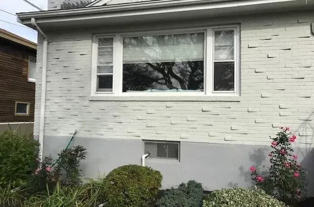 A white brick house exterior with a large window, flanked by two smaller windows. Below, a garden with various plants and pink flowering bushes.