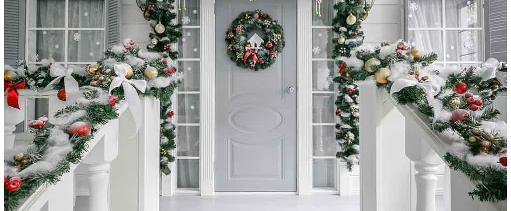 A festive porch features a gray door adorned with a holiday wreath. Lush garlands with red and gold ornaments decorate the railing, creating a cozy, welcoming atmosphere.