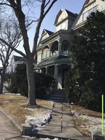 Victorian-style house in winter, with intricate woodwork, a porch, and steep gabled roof. Bare trees and patches of snow line the sidewalk.