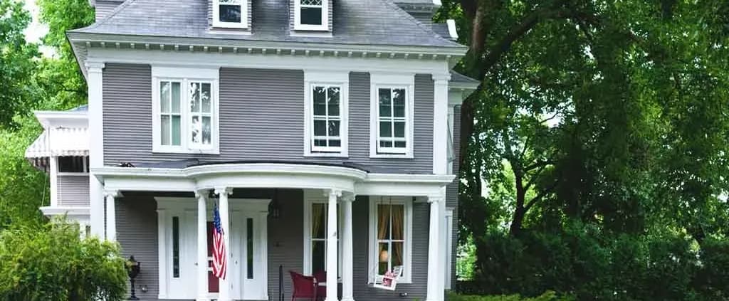 Gray Victorian-style house with white trim, featuring a porch, columns, American flag, and surrounded by lush green trees, conveying a serene, classic feel.
