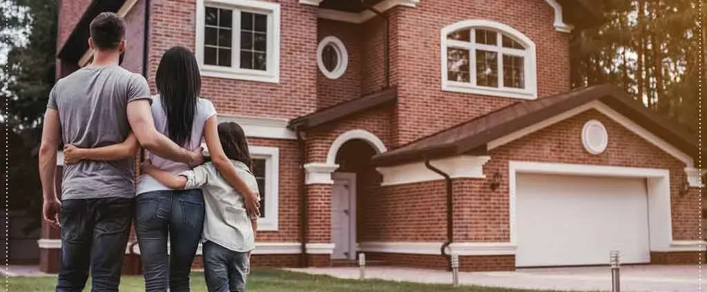 A family of three stands with arms around each other, facing a large red brick house with white accents, conveying a sense of togetherness and new beginnings.