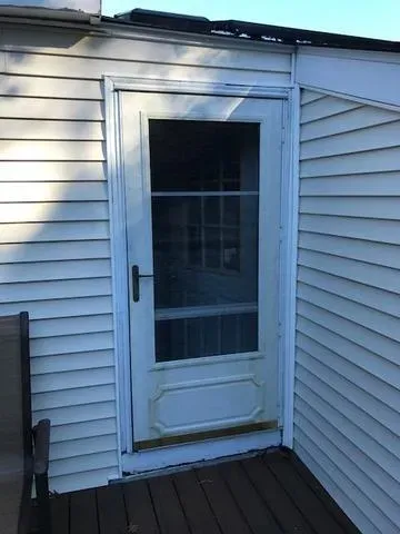 A white storm door opens into a solid wall of siding on a house exterior, creating a humorous and confusing architectural mistake. Shadowed and bright tones.