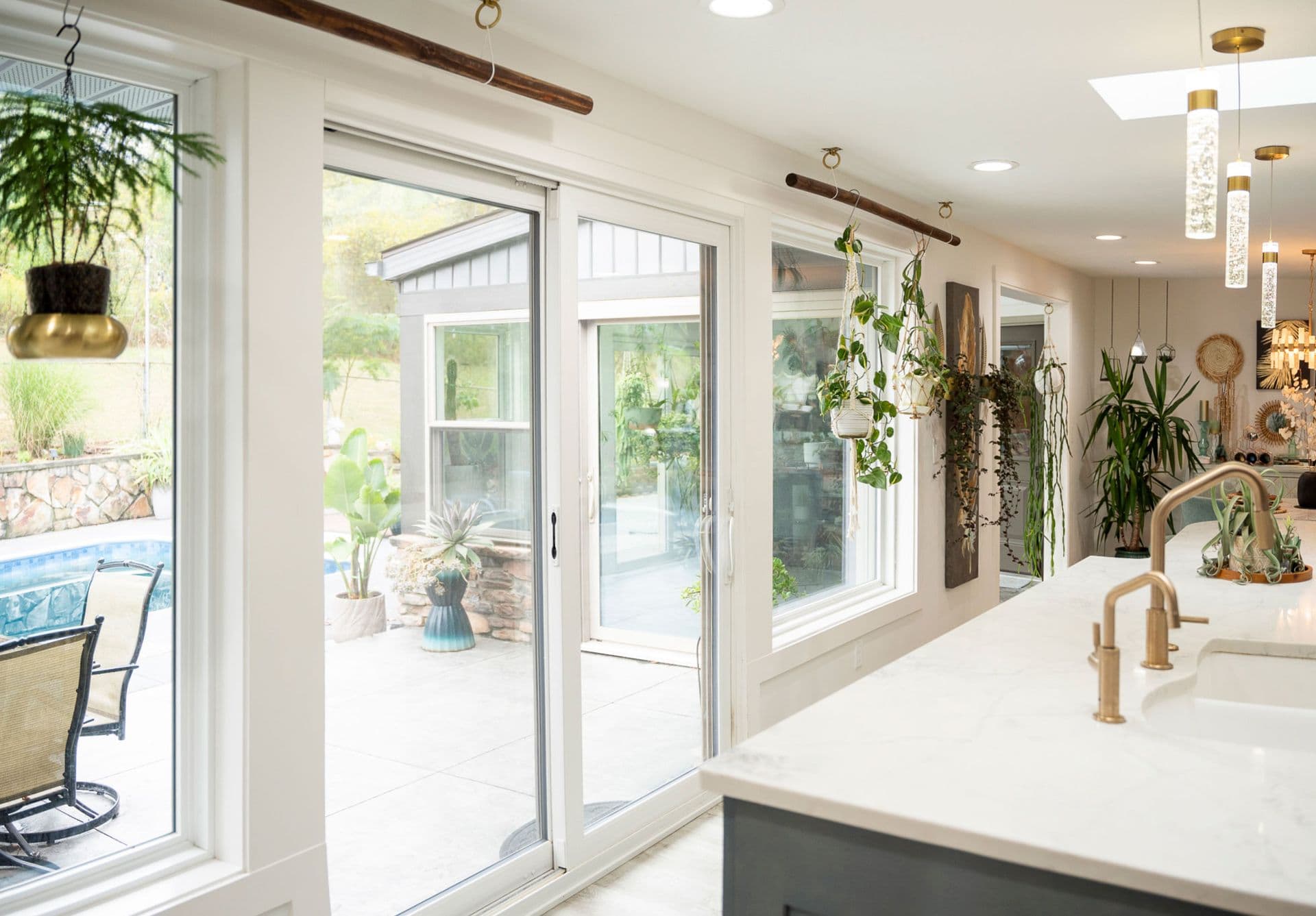 Modern kitchen with white countertops, gold fixtures, and hanging plants. Large glass doors open to a patio with pool, exuding a fresh, airy vibe.