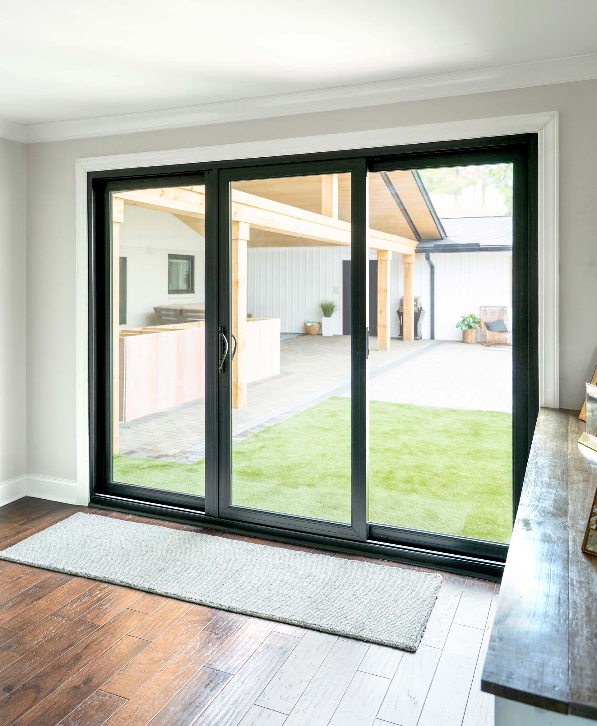 Sliding glass doors open to a patio with green grass and wooden overhangs. Sunlight illuminates the spacious, modern interior with hardwood floors.
