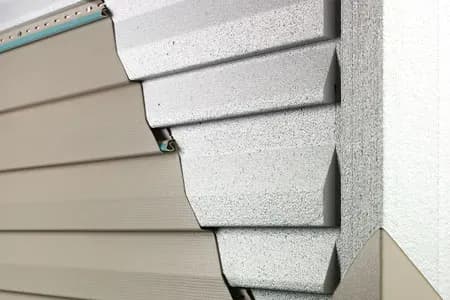 Close-up of a house corner showing layers of beige vinyl siding beneath overlapping gray stucco panels. The mix of textures gives a modern, clean look.