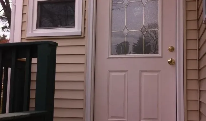 A beige front door with decorative frosted glass panels is set in a house with beige siding. A small window is on the left, above a green railing.