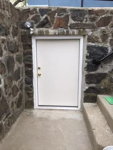 Small white door set in a stone wall, with dual handles, and next to concrete steps leading upwards. The scene feels mysterious and quaint.
