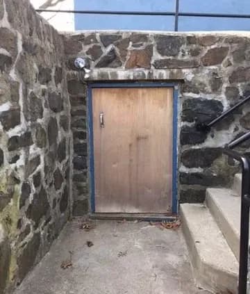 A small, wooden door set in a stone wall beside concrete steps. The scene is outdoors, with fallen leaves on the ground, conveying a sense of mystery.