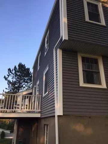 Three-story house with gray siding and white trim, viewed from below. A deck and trees are visible, and the scene is lit by the late afternoon sun.