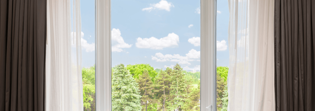 Wide window view with sheer white curtains framing a lush green forest under a clear blue sky with fluffy clouds; serene and inviting.