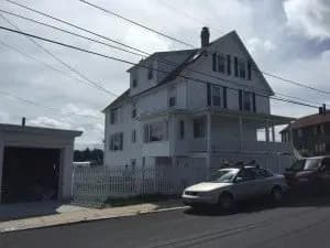 A large, white, three-story house with a porch and picket fence on a cloudy day. A car and an SUV are parked on the street next to the house.