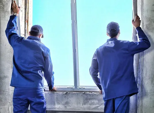 Two workers in blue uniforms and hats install a large window in a building under construction. The scene conveys teamwork and construction progress.