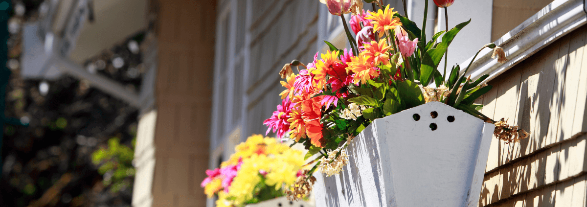 White window boxes filled with vibrant pink, yellow, and orange flowers are attached to a beige house. The scene is bright and cheerful.