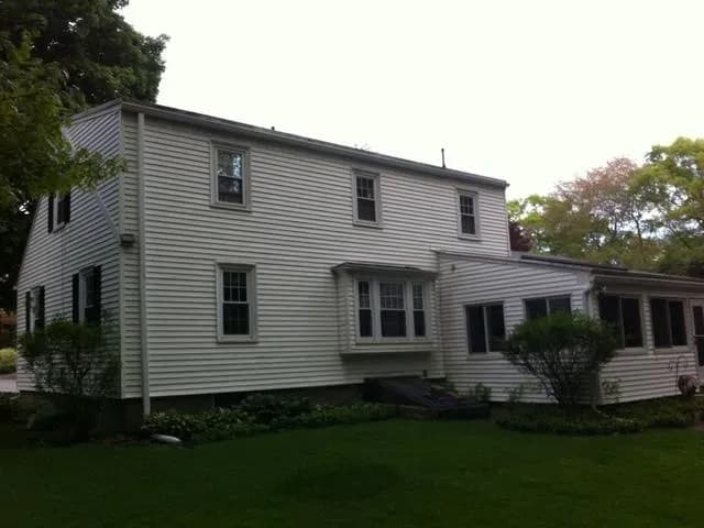 Two-story white wooden house with multiple windows and a small porch, surrounded by green lawn and trees. The atmosphere is calm and residential.