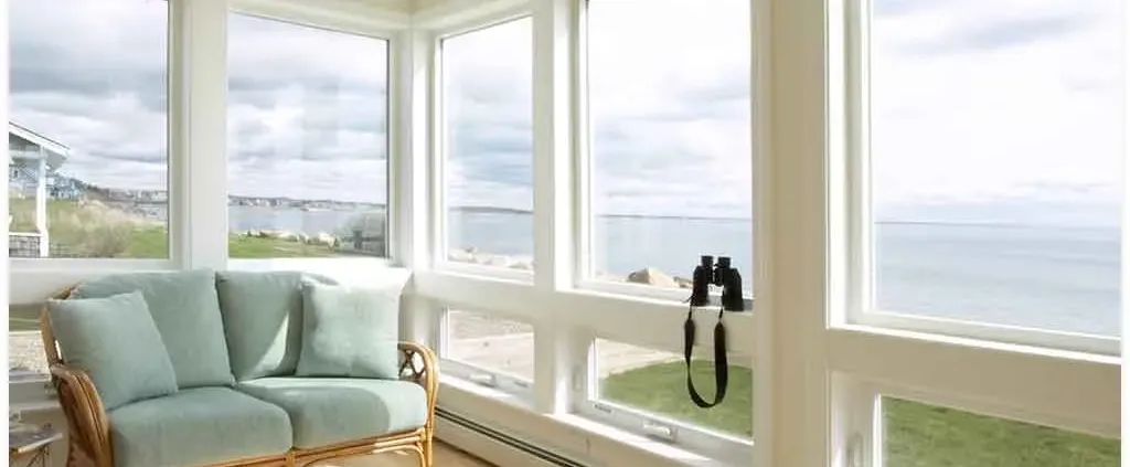 Bright sunroom with large windows overlooking the ocean, showcasing a scenic view. A cozy rattan loveseat with green cushions adds a relaxing feel.