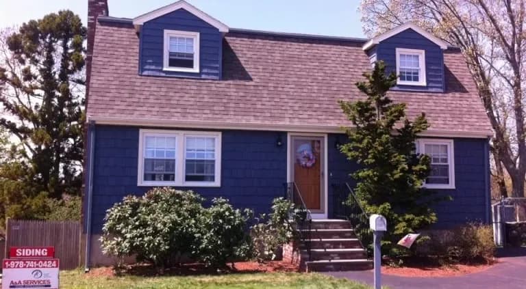 A charming blue two-story house with a brown shingled roof, white-trimmed windows, a front door with a wreath, and a manicured lawn with shrubs.