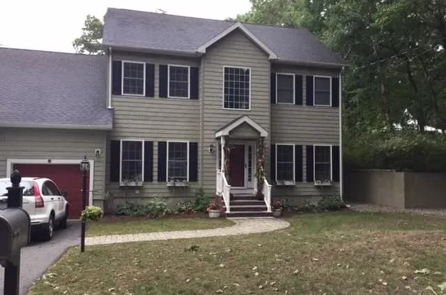 Two-story gray house with black shutters, situated in a lawn with a stone path. A white SUV is parked in the driveway beside a black mailbox.