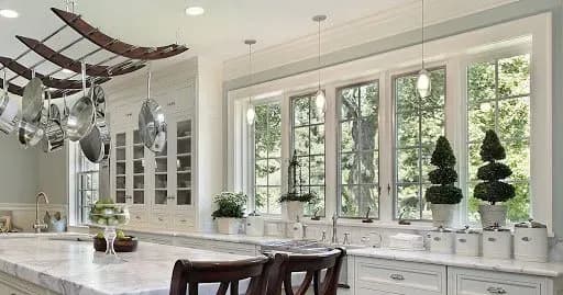 Spacious kitchen with large windows, white cabinets, and marble countertops. Hanging pots, potted plants, and cozy lighting create an elegant ambiance.