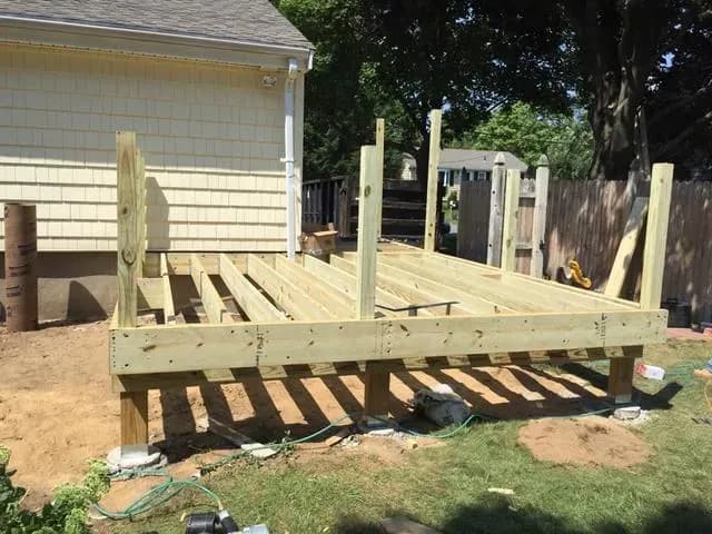 Wooden deck under construction beside a house, featuring exposed beams and posts. Nearby, a sunny yard and garden hose are visible.