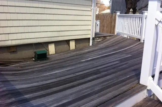 Warped wooden deck with wavy, uneven boards, adjacent to a house with beige siding. White railings show bending, suggesting structural damage.