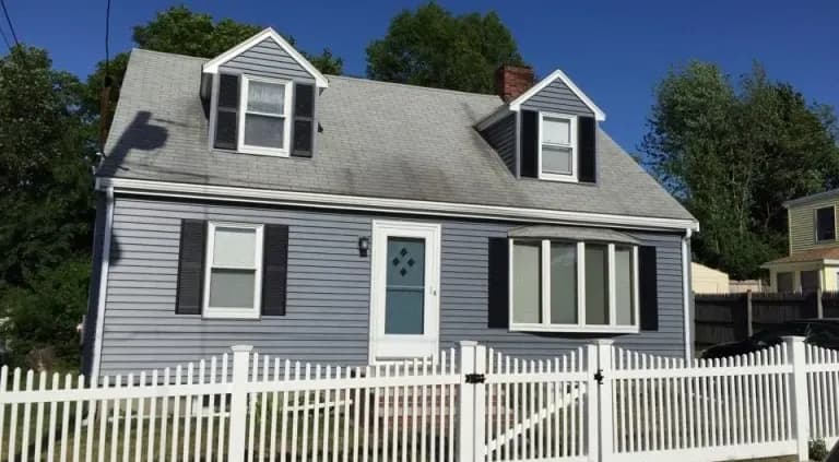 A gray, two-story house with white trim and blue door, featuring dormer windows and dark shutters, is surrounded by a white picket fence on a sunny day.