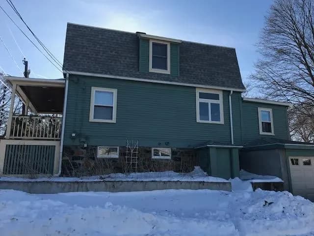 Two-story blue house with a covered porch and a snowy yard. It has a steep roof, several windows, and a garage, creating a cold, wintery tone.