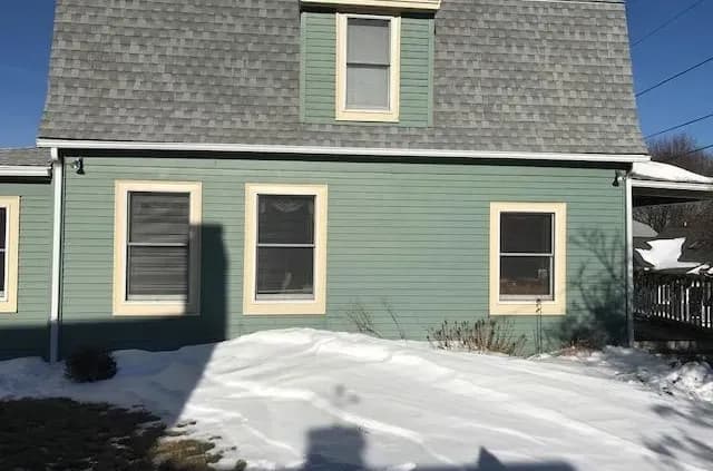 A light green house with a gambrel roof and snow-covered yard. Three windows on the sunny side, casting shadows. Clear blue sky above.