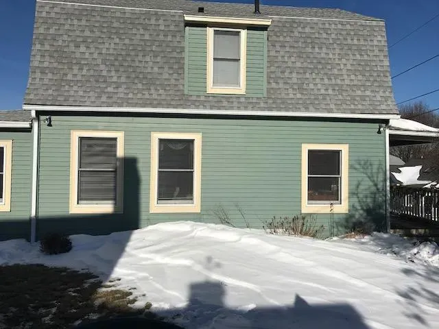 Green house with a gray roof in winter; snow covers the ground. The home has beige window frames and a dormer window. Bright, clear sky.