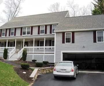 Two-story gray house with white trim, large porch, and maroon shutters. A white car is parked in the driveway. Lawn and bare trees in the background.