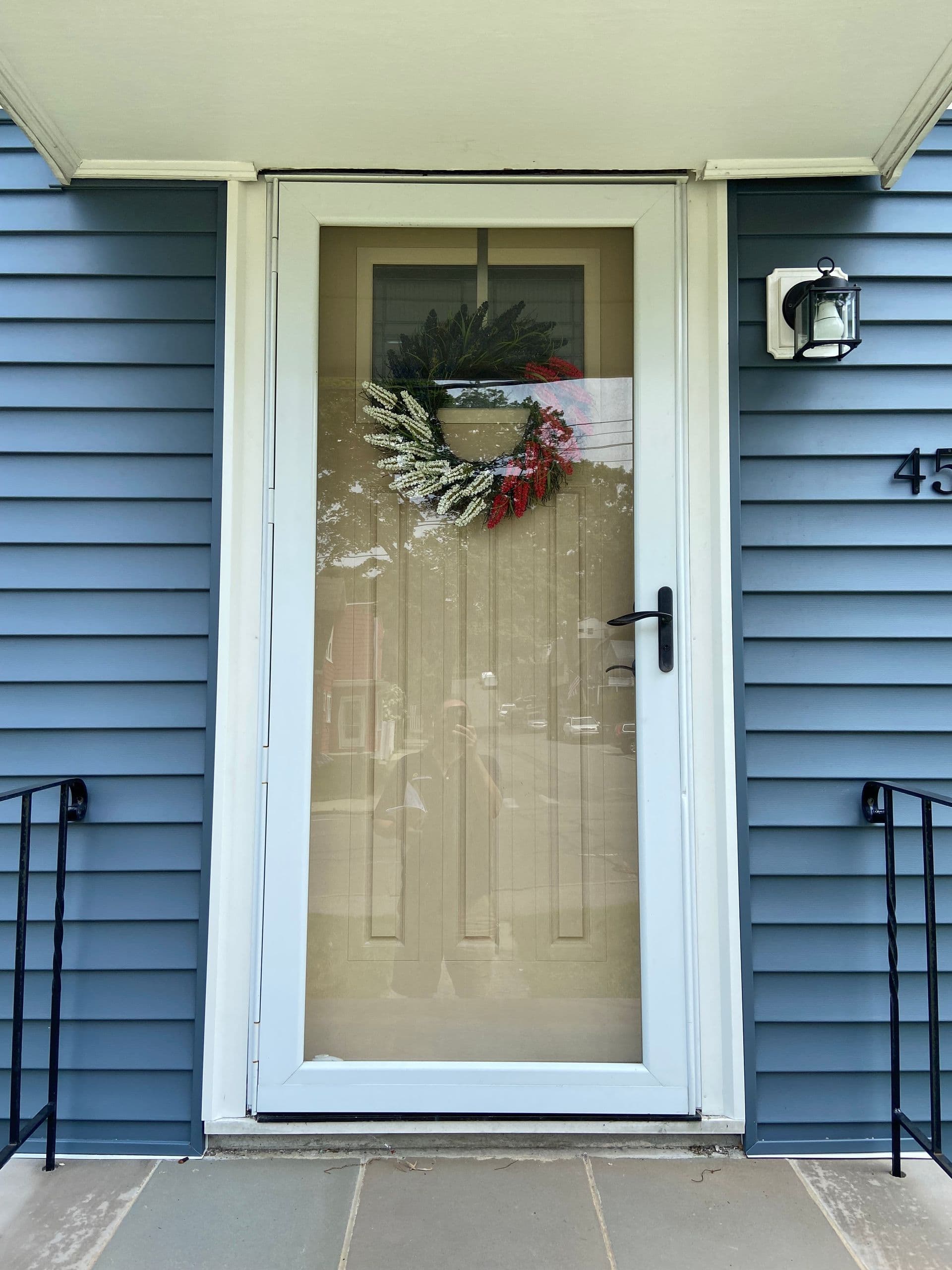 A blue house exterior features a white front door with a glass storm door. A festive wreath with red and white accents decorates the door, creating a welcoming and cheerful entry.