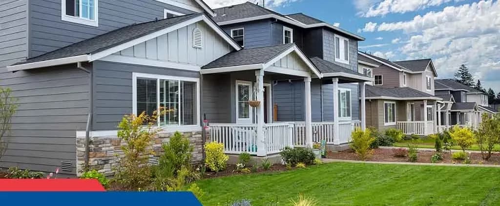 A row of modern suburban homes with gray siding and front porches, manicured lawns, and small shrubs. The sky is partly cloudy, creating a peaceful suburban atmosphere.