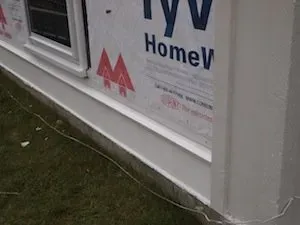 Close-up of a house exterior under construction, showing a window with white trim, Tyvek house wrap, and grass below. The scene suggests ongoing renovations.