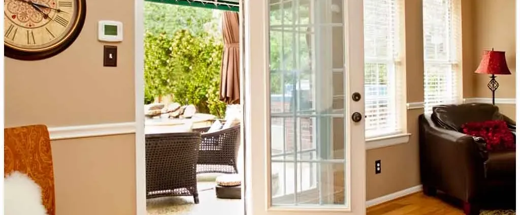 Sunlit living room with a glass door leading to a patio. Inside, a brown armchair and red lamp create a cozy feel. Outside, wicker patio furniture is visible.