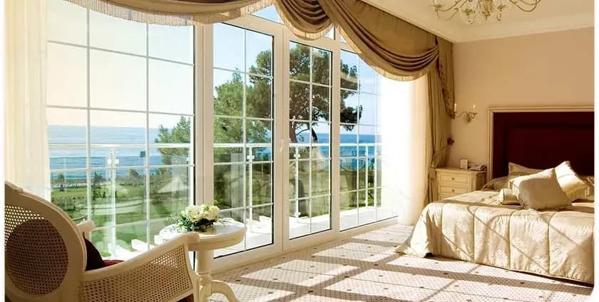 Luxurious bedroom with a large window showcasing a seaside view. Sunlight bathes the elegant decor, featuring a gold bedspread and plush furniture.