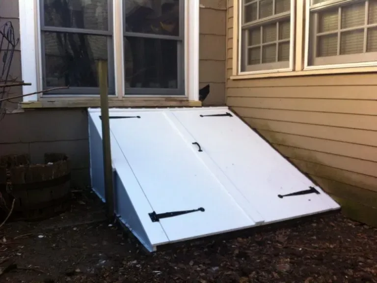 White cellar doors are installed vertically over a window on a tan house, surrounded by dirt and a barrel planter, creating an unconventional setup.