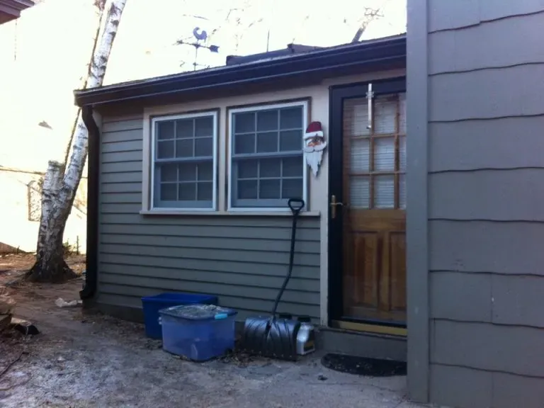 Small gray house with two windows and a wooden door on a sunny day. A Santa decoration hangs on the door, and a shovel is propped outside.