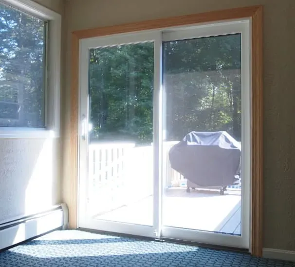 Sunlit room with sliding glass doors leading to a deck with a grill. Bright daylight creates a serene and inviting atmosphere, highlighting the greenery outside.