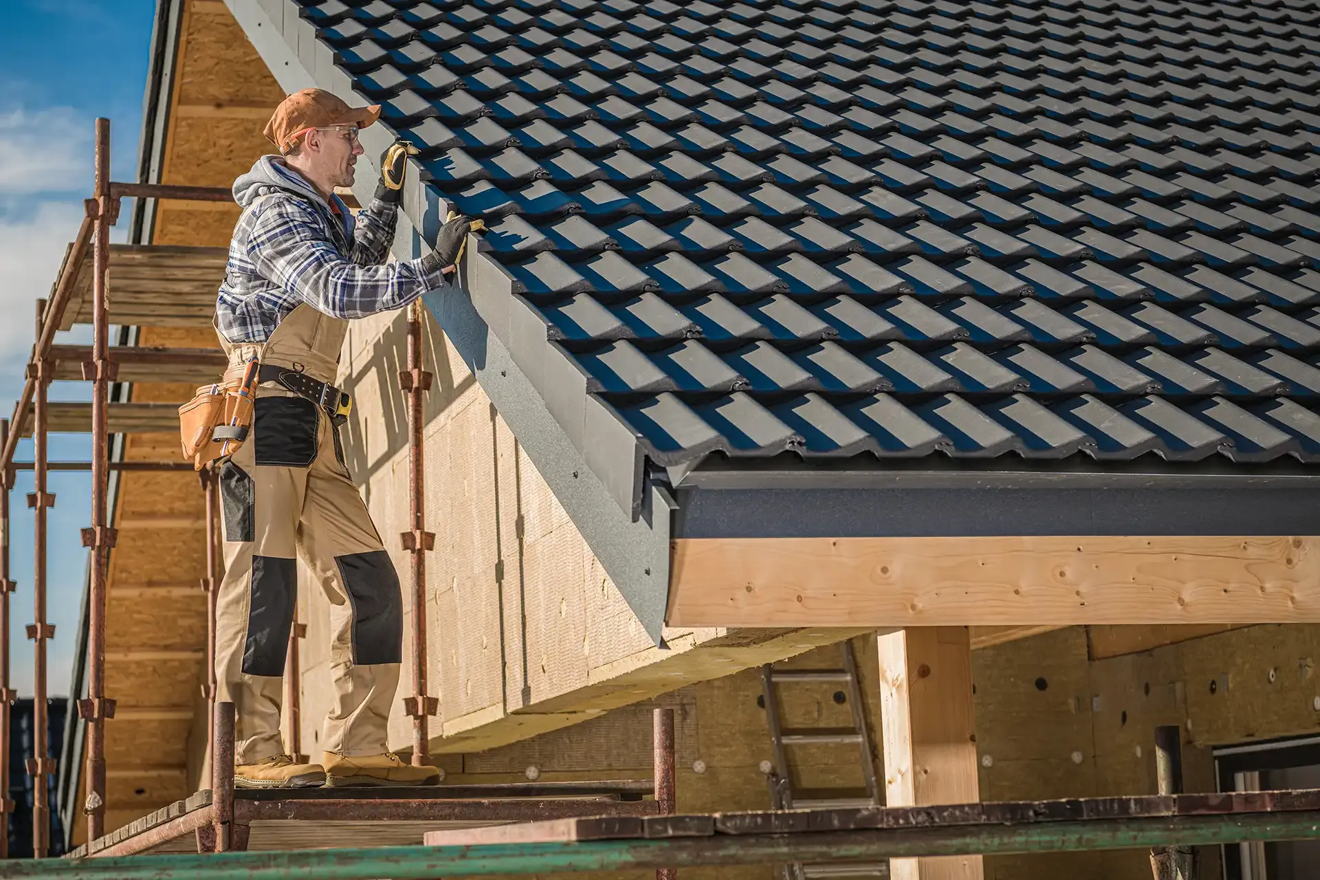 Roofer on scaffolding installs black metal tiles. The worker wears safety gear and a tool belt, set against a clear sky and wooden structure.