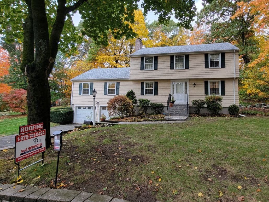 Two-story beige house with black shutters surrounded by autumn trees. A "Roofing" sign is on the lawn, indicating repair services. Overcast sky.