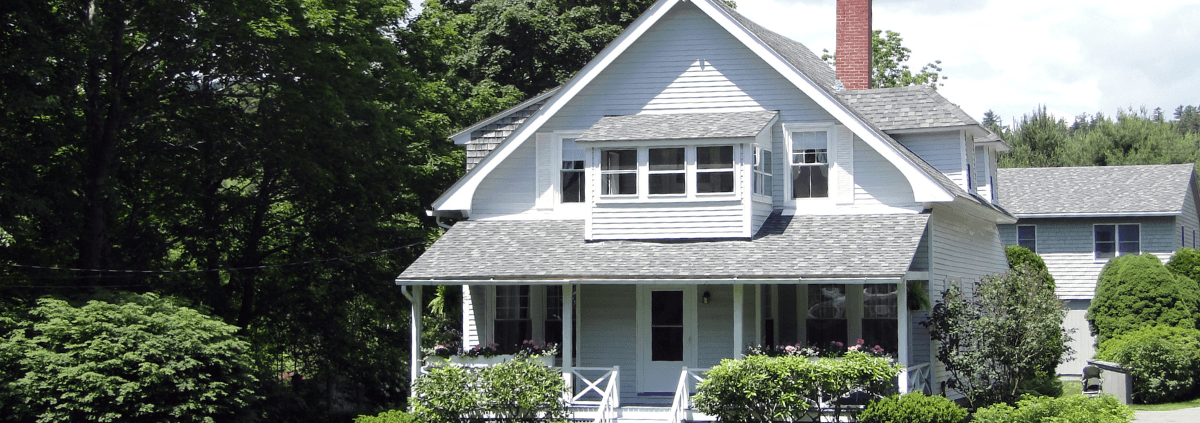 Charming white house with gray roof, featuring double-tiered windows and a porch. Surrounded by lush greenery and a sunny sky, conveying a peaceful, inviting atmosphere.