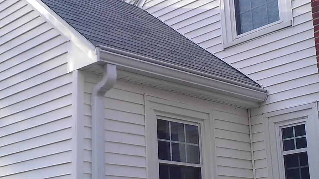 A white house corner with gray sloped roof, seamless gutter system, and downspout. Two windows with grid patterns. Clean, neat appearance.