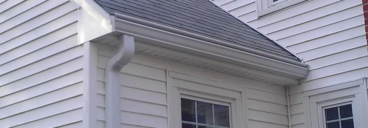 A white house corner with vinyl siding, featuring gray roof shingles, a white downspout, and two windows below, conveying a clean, well-maintained look.