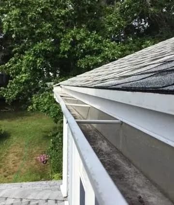 Rooftop gutter with shingle roof, viewed from above. The gutter appears clean, bordered by trees and grass, creating a peaceful outdoor setting.